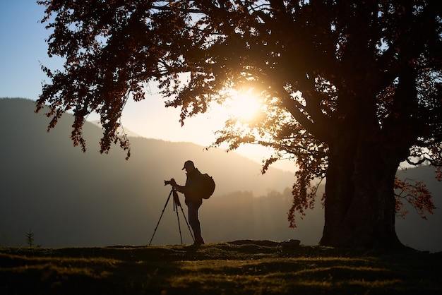 Homem de turista caminhante com câmera no vale gramado da paisagem montanhosa sob uma árvore grande.