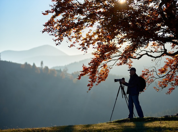 Homem de turista caminhante com câmera no vale gramado no fundo da paisagem montanhosa sob uma árvore grande.