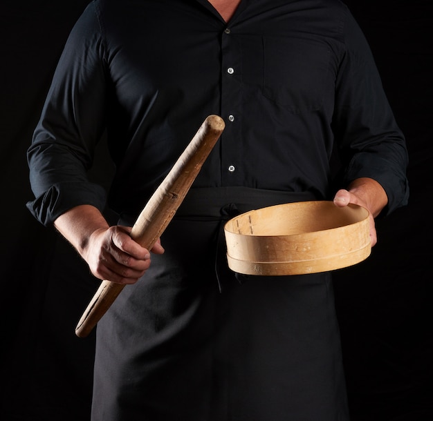 Homem de uniforme preto, segurando a peneira de madeira redonda vintage vazia para peneirar a farinha e o rolo, chef fica contra o fundo preto