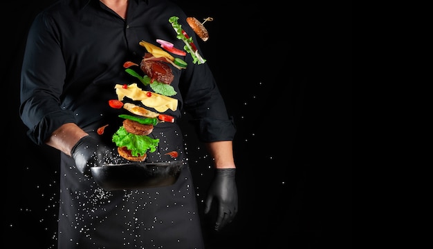 Homem de uniforme preto segurando uma frigideira redonda de ferro fundido com ingredientes de um cheeseburger levitando: pão de gergelim, queijo, tomate, cebola, costeleta de carne, pimenta, espaço de cópia