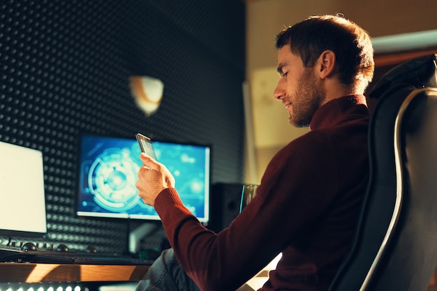 Homem de vista lateral sentado em uma cadeira de couro, trabalhando no estúdio, usando um smartphone e computadores. Freelancer tem celular trabalhando em filmagens, vídeos, design.