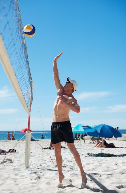 Homem de vôlei e fitness na praia com bola serve energia ou liberdade em fundo de céu azul Esportes de praia ou jogador masculino com handebol na natureza para treino de cardio ou desempenho de jogo atlético
