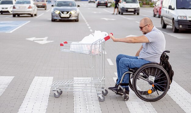 Foto homem deficiente em cadeira de rodas empurrando carrinho na frente de si no estacionamento de um supermercado