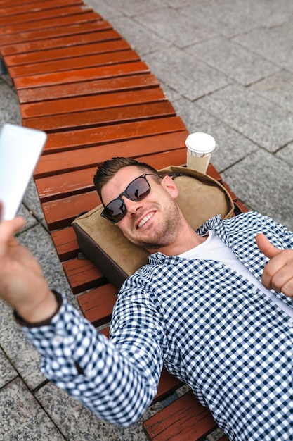 Homem deitado em um banco de parque fazendo selfie com celular
