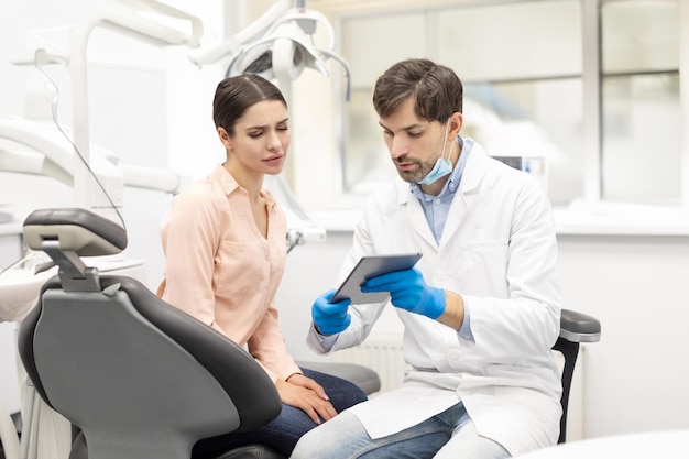 Homem dentista em roupa branca mostrando tablet digital para jovem cliente mulher sentada em uma cadeira em