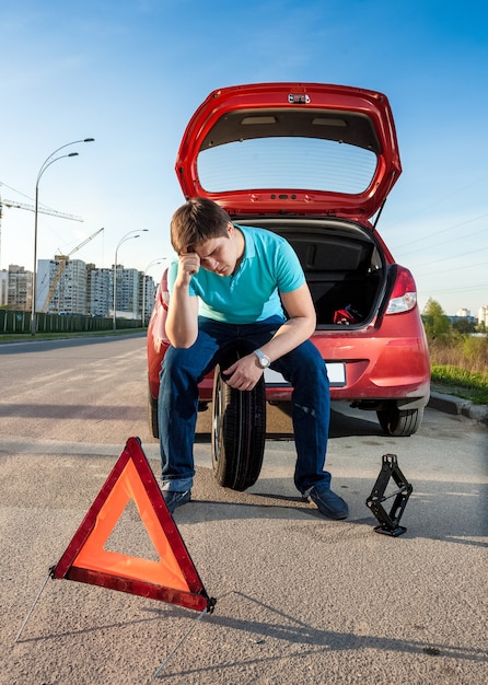 Homem deprimido sentado no estepe perto de um carro quebrado