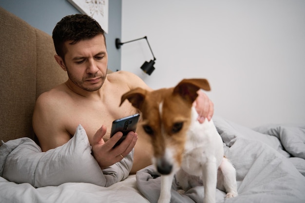 Homem descansando na cama com cachorro e usa smarpthone