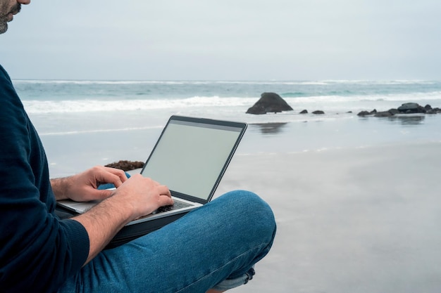 Homem desconhecido sentado relaxado na praia com um laptop trabalhando com vista para as mãos