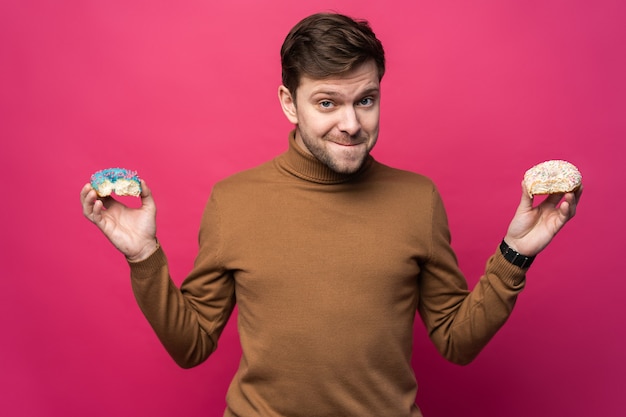 Homem descontente segura dois saborosos donuts de anel, sente-se infeliz porque não consigo comer doces, isolados sobre o fundo rosa.