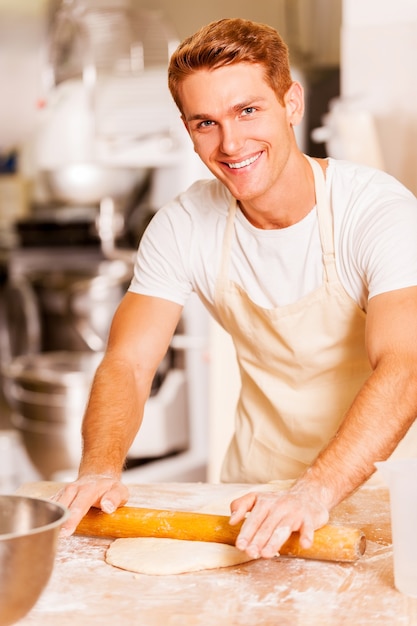 Homem desenrolando a massa. Jovem e bonito padeiro sorridente abrindo a massa com um rolo de massa