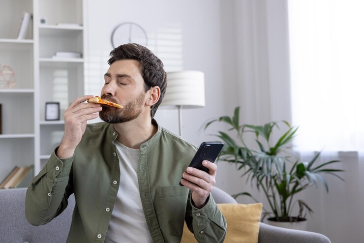 Foto homem desfrutando de uma fatia de pizza enquanto usa smartphone em casa