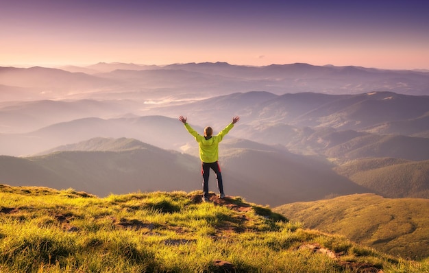 Homem desportivo de pé no pico da montanha com os braços levantados contra o vale da montanha no nevoeiro ao pôr do sol no outono Jovem feliz balança a floresta e o céu azul no outono Viajante caminhando nas montanhas