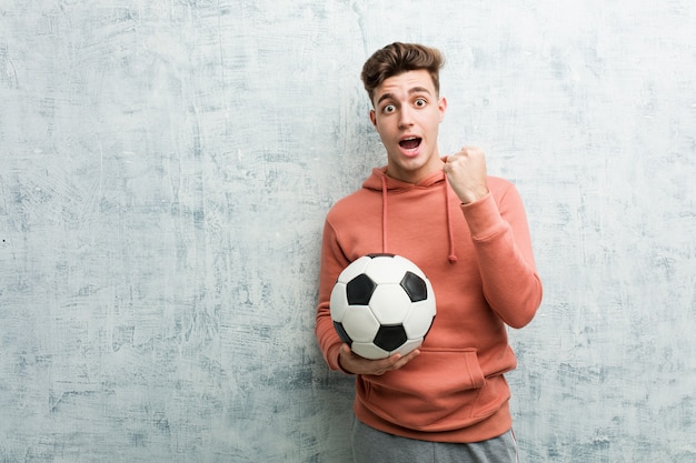 Homem desportivo novo que guarda uma bola de futebol que cheering despreocupado e entusiasmado. Conceito de vitória.