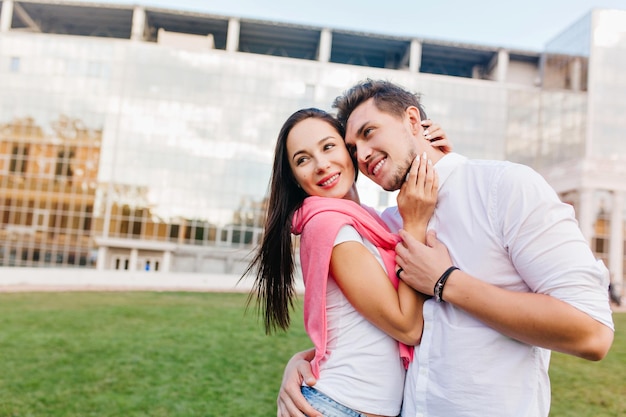 Homem desportivo usa pulseira preta, aproveitando o tempo livre com uma garota charmosa e sorridente. Mulher de cabelos escuros incrível, abraçando-se com o marido na sessão de fotos ao ar livre na cidade.