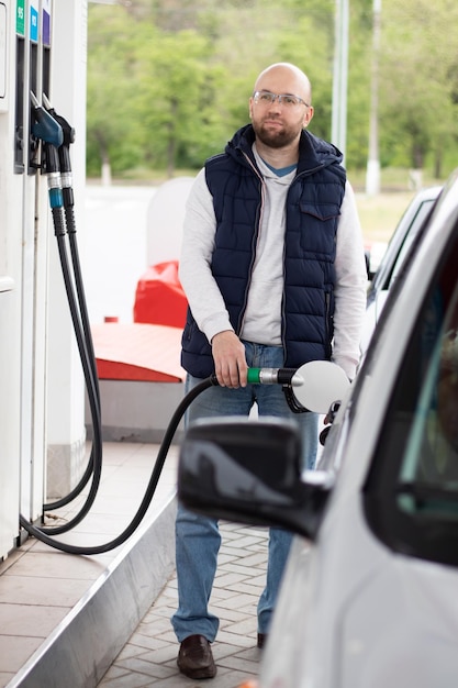 Homem do milênio vestido com jeans e um colete escuro reabastece seu carro prateado em um posto de gasolina