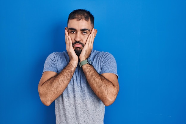 Homem do oriente médio com barba em pé sobre fundo azul mãos cansadas cobrindo rosto depressão e tristeza chateado e irritado por problema
