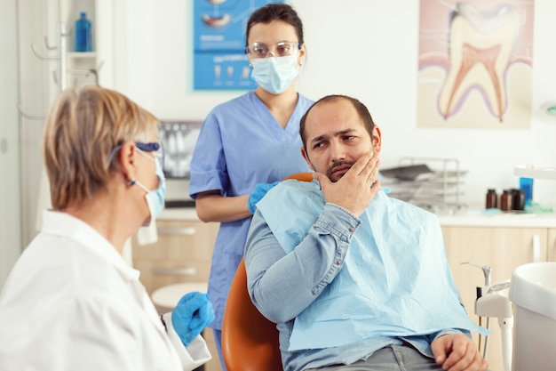 Homem doente com sintomas de dor de dente, segurando a mão na bochecha enquanto fala com o médico sênior