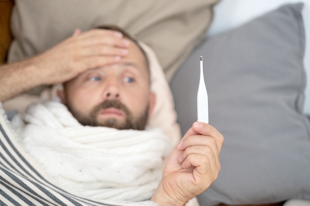 Homem doente, deitado no sofá, verificando sua temperatura em casa, na sala de estar