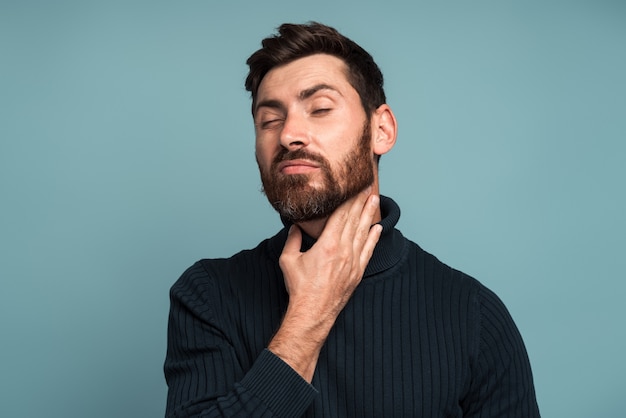 Homem doente infeliz tocando o pescoço, sentindo dor ao engolir, resultado de calafrios, inflamação da garganta, dor de garganta. Foto de estúdio interna isolada em fundo azul