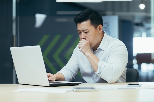 Homem doente no escritório tosse e tem um empresário asiático de nariz escorrendo com trabalhos frios no escritório no laptop