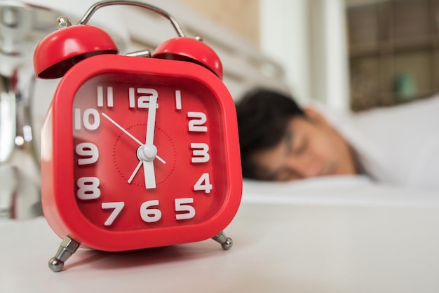 Homem dormindo na cama com despertador vermelho em primeiro plano em casa