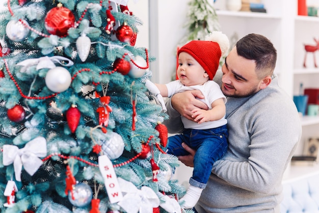 Homem e bebê fofo estão prontos para comemorar o ano novo. feliz pai brinca  com seu filho bebê perto de árvore de natal na noite de natal. | Foto  Premium