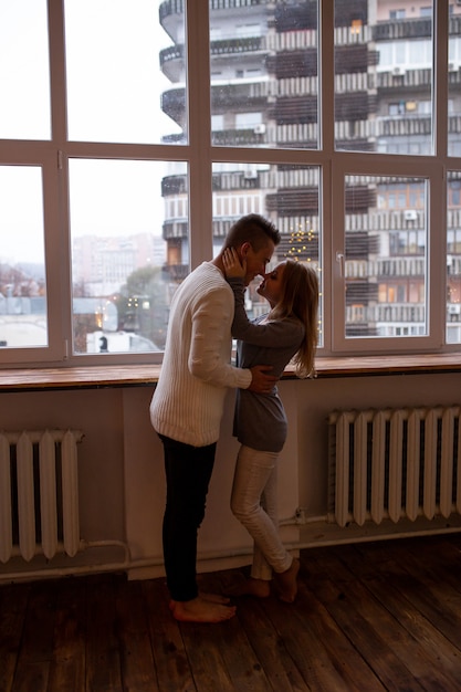 Homem e mulher apaixonada se abraçam em pé perto da janela em casa