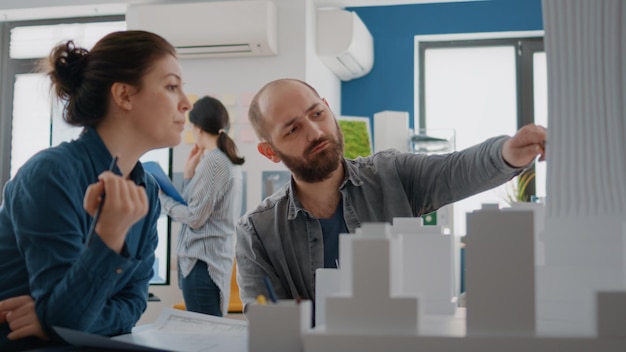 Foto homem e mulher apontando para o modelo de construção para planejar projetos no papel. colegas usando maquete para projetar estrutura de construção, auxiliando no desenvolvimento arquitetônico. projeto industrial