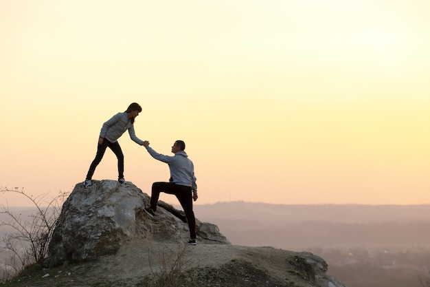 Foto homem e mulher caminhantes ajudando uns aos outros para escalar pedra ao pôr do sol nas montanhas