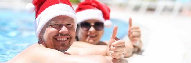 Homem e mulher com chapéu de Papai Noel descansando na beira da piscina e mostrando o polegar