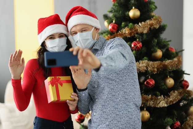 Homem e mulher com chapéu de Papai Noel e máscaras protetoras nos rostos estão segurando um telefone celular e acenando perto