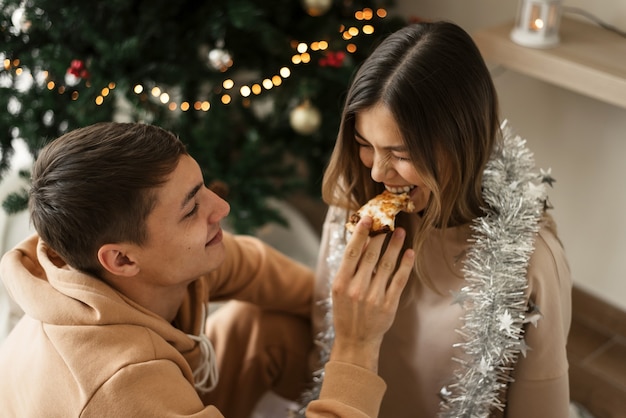 Homem e mulher comendo pizza em casa perto da árvore de natal e se divertindo