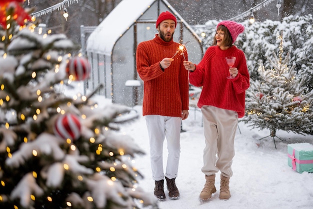 Homem e mulher de suéteres vermelhos celebram as férias de Ano Novo acendendo faíscas e se divertindo perto da árvore de Natal no quintal nevado