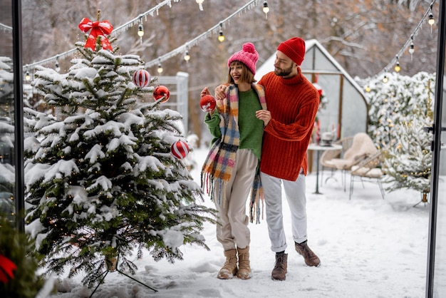 Homem e mulher decoram a árvore de Natal com bolas festivas enquanto se preparam para as férias de inverno no quintal nevado de sua casa Família feliz celebrando as férias de Ano Novo