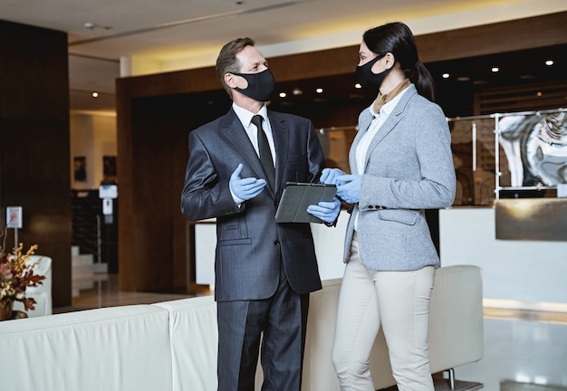 Homem e mulher elegantes com máscaras médicas e luvas de borracha olhando um para o outro enquanto se comunicam no saguão de um hotel