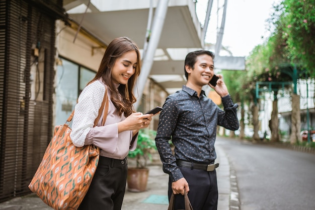 Homem e mulher em pé na calçada, sorrindo, usando o telefone