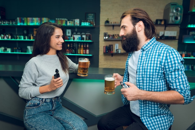 Homem e mulher estão sentados com cerveja e cigarro eletrônico.