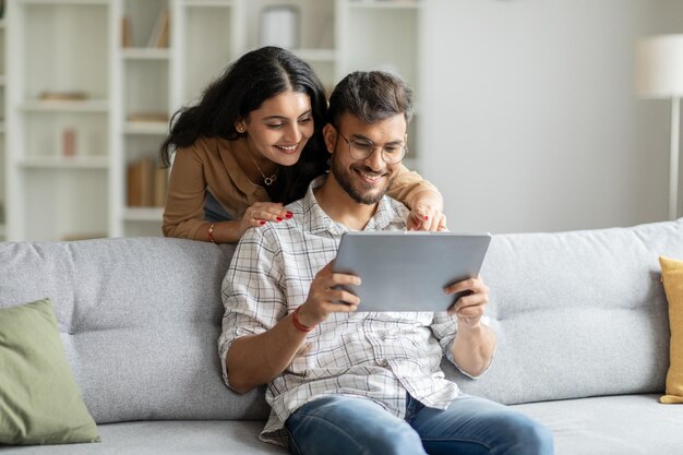 Homem e mulher indianos felizes sentados no sofá abraçando-se e usando tablet digital olhando para a tela do pad