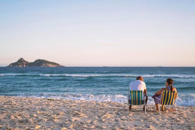 Homem e mulher relaxam em cadeiras listradas dobráveis em uma praia arenosa em frente ao oceano ao pôr do sol