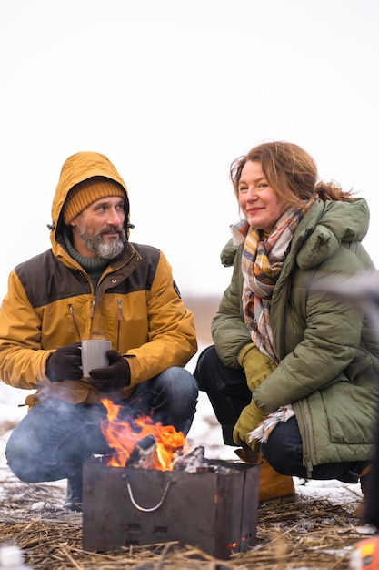 Homem e mulher se aquecendo perto do fogo na natureza bebendo chá quente ou café Casal maduro fazendo um churrasco na floresta de inverno na fogueira Conceito de viagem em família Conceito de férias em família