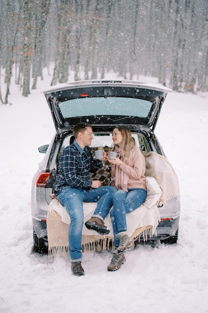 Homem e mulher sorridente com copos nas mãos estão sentados no porta-malas de um carro em uma floresta coberta de neve