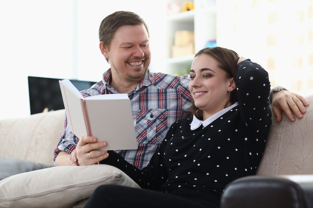 Homem e mulher sorrindo estão sentados no sofá segurando um livro