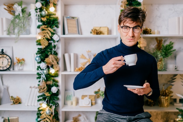 Homem elegante com rosto emocional positivo, posando para a câmera com uma xícara de café na sala aconchegante com árvore cristmas e decorações de ano novo