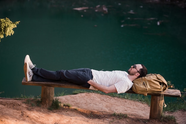 Homem elegante de alpinista, deitado num banco de madeira perto do lago