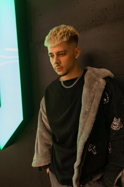 Homem elegante jovem hippie com penteado com uma roupa preta da moda com uma jaqueta da moda e camiseta preta fica perto de uma parede preta com uma luz verde interna