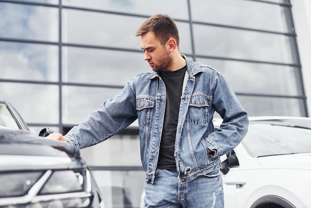 Homem elegante parado perto de carro ao ar livre contra o edifício empresarial moderno