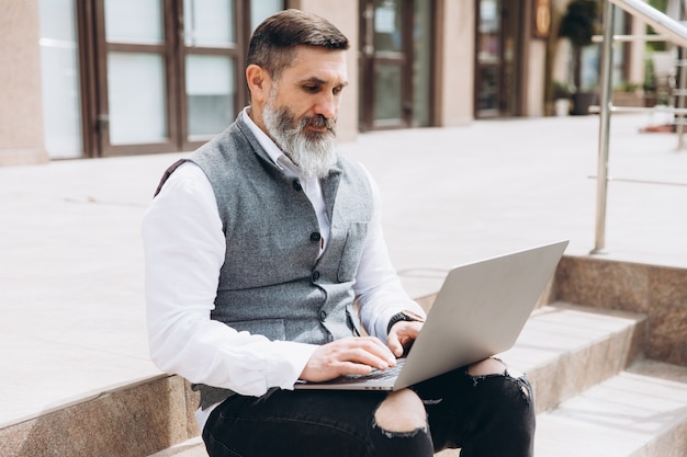 Homem elegante sênior com barba grisalha passa o tempo ao ar livre no laptop.