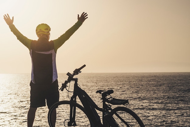 Homem em frente ao mar com os braços erguidos, olhando para o horizonte ao pôr do sol com sua bicicleta - conceito de estilo de vida de liberdade e piloto sênior