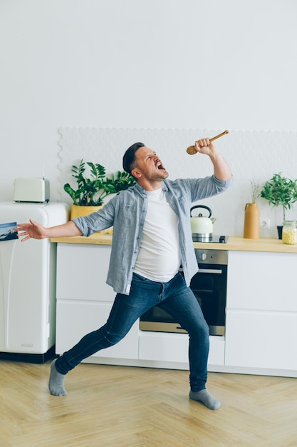 Homem em roupas casuais cantando na colher na cozinha dançando curtindo a vida