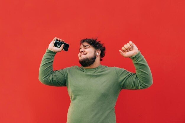 homem em roupas casuais fica em um fundo vermelho com um smartphone na mão e ouve música